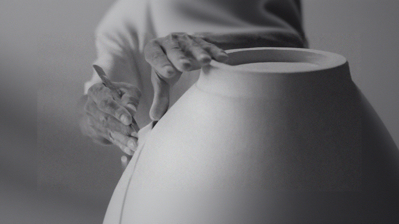 Person holding a large ceramic vase with a neutral background, trimming a pottery piece on the wheel 